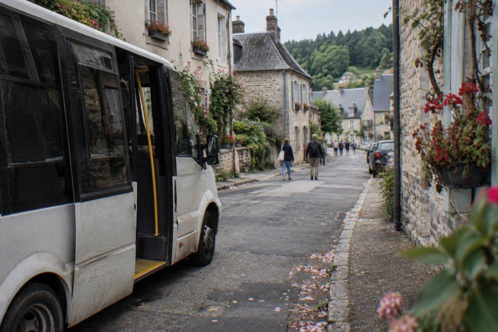 A small shuttle bus parked on a village street lined with stone houses and flower beds