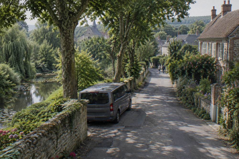 A minivan parked on a tree-lined lane near a stone garden wall in morning light