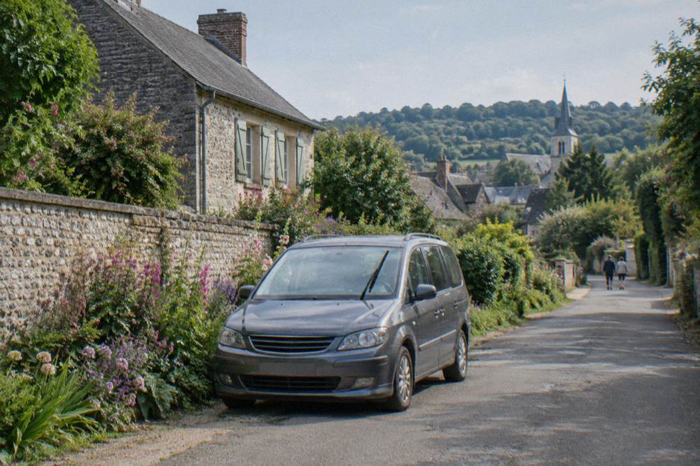 A small minivan parked on a country lane beside flowering gardens at the edge of a village