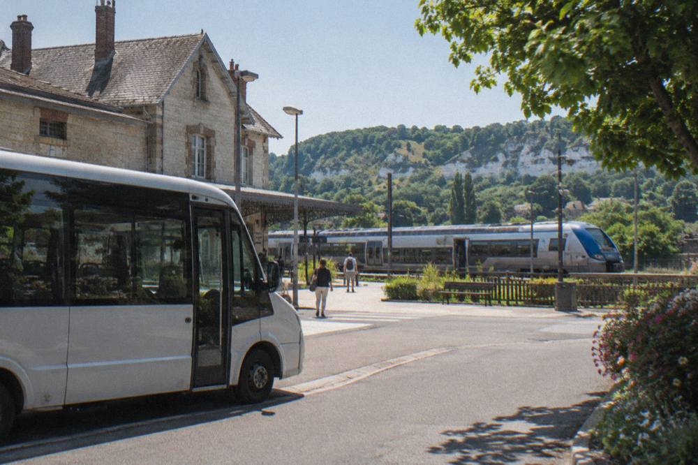 A regional train standing at a small Normandy station platform with a shuttle bus waiting outside