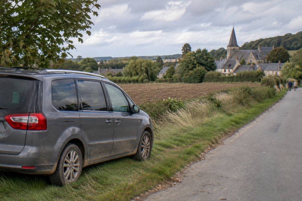 A tour minivan parked on a lane near a Normandy village with hedgerows and fields beyond