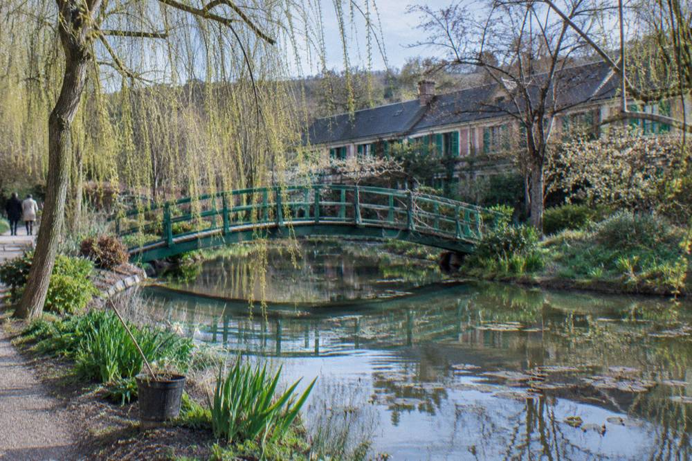 Arched green footbridge over a pond with water lilies and willow trees